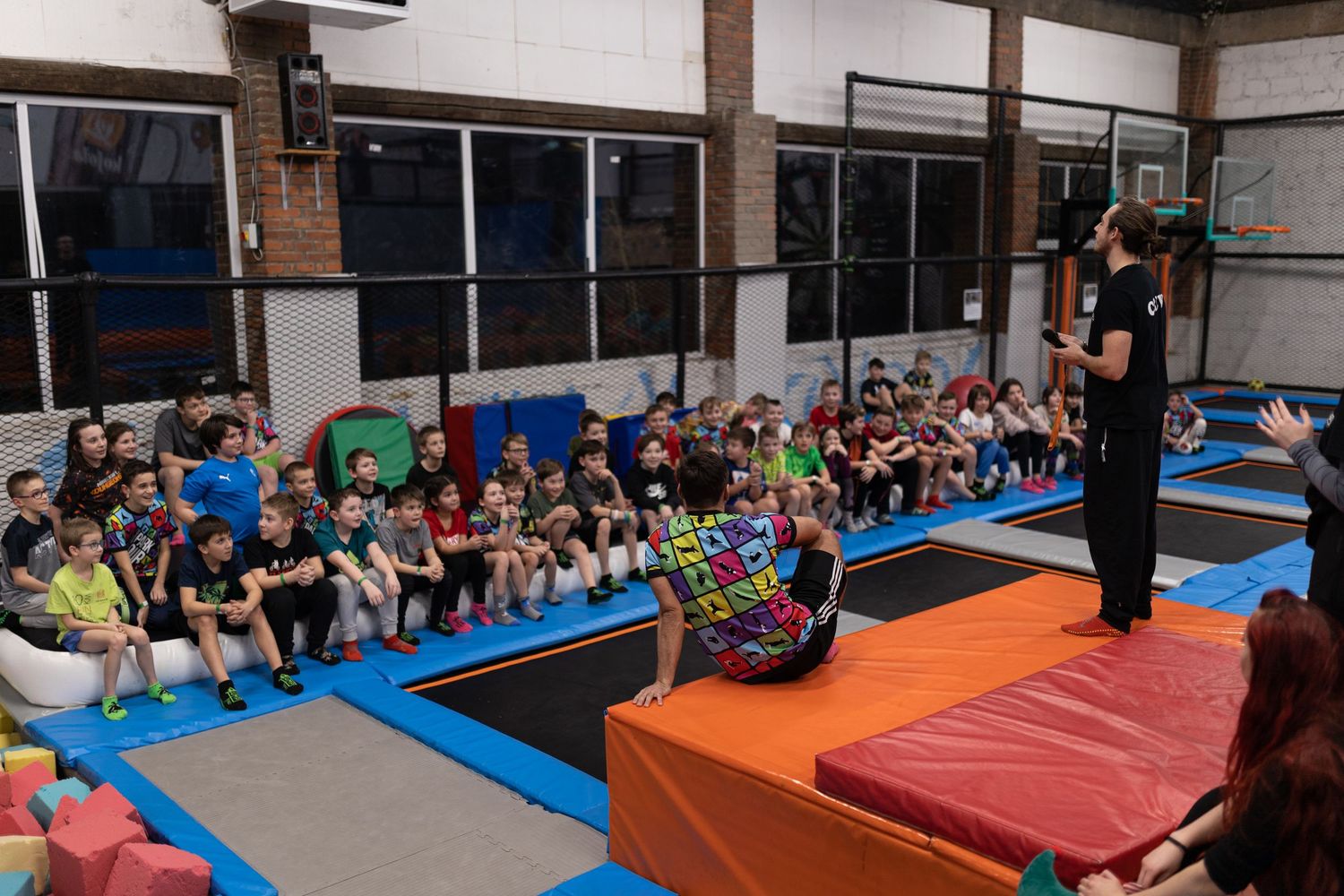 A group of people sit along the wall watching others perform exercises at a gymnastics or fitness facility.