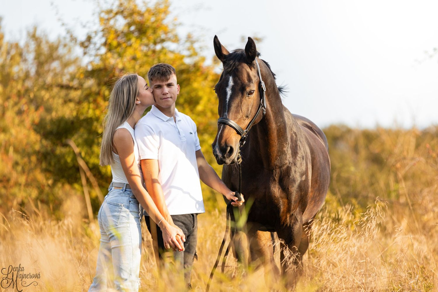 A bay horse stands with people in a golden autumn field.