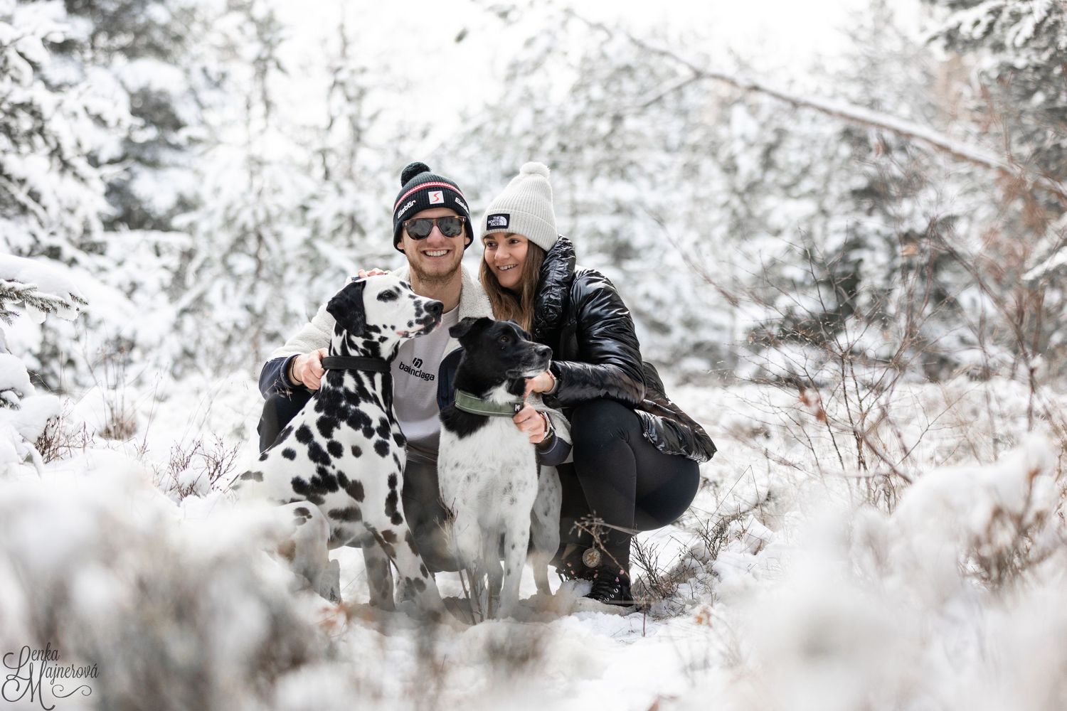Two people pose with their black and white dog in a snowy winter forest setting.