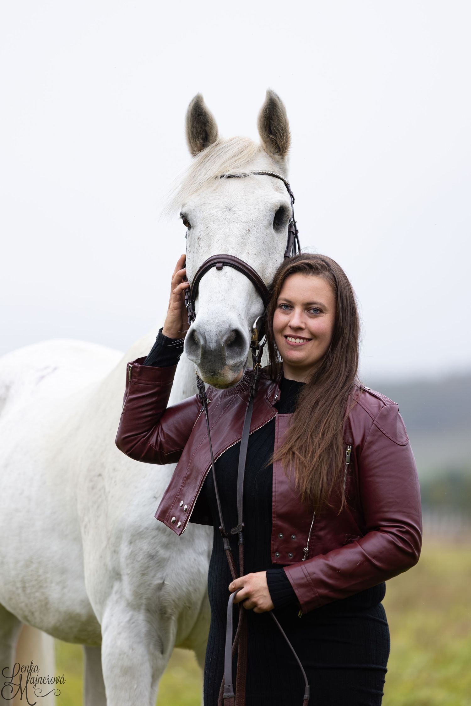 A white horse stands with its handler wearing a burgundy leather jacket in a misty outdoor setting.