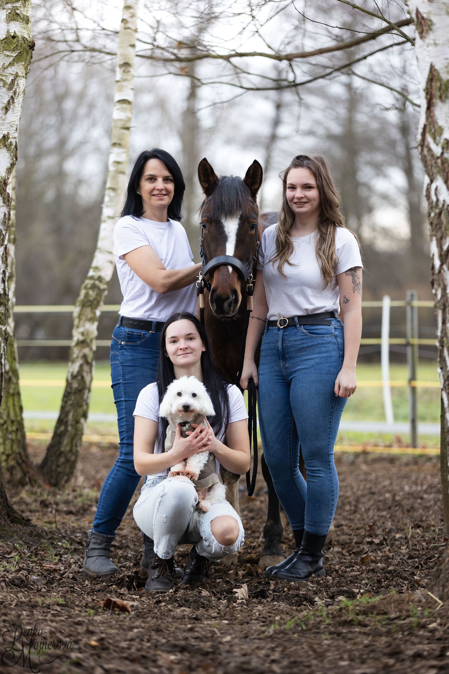 A group poses outdoors with a black horse and white dog among trees on a wooded path.