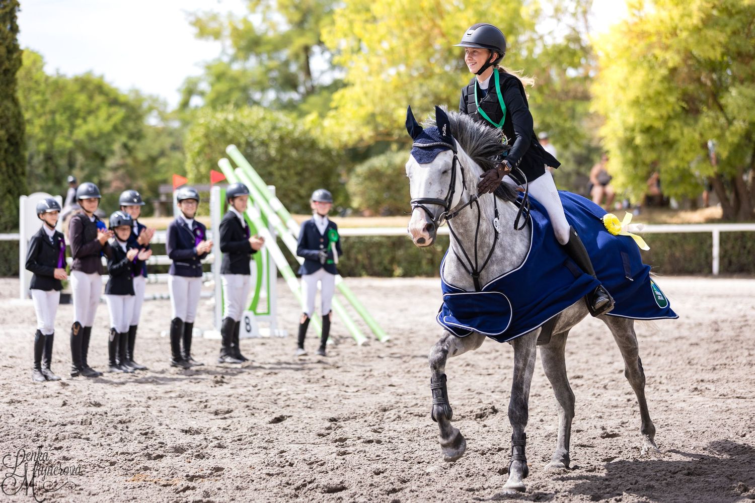 An equestrian rider in blue attire performs on a gray horse while spectators in white uniforms watch from the sidelines.