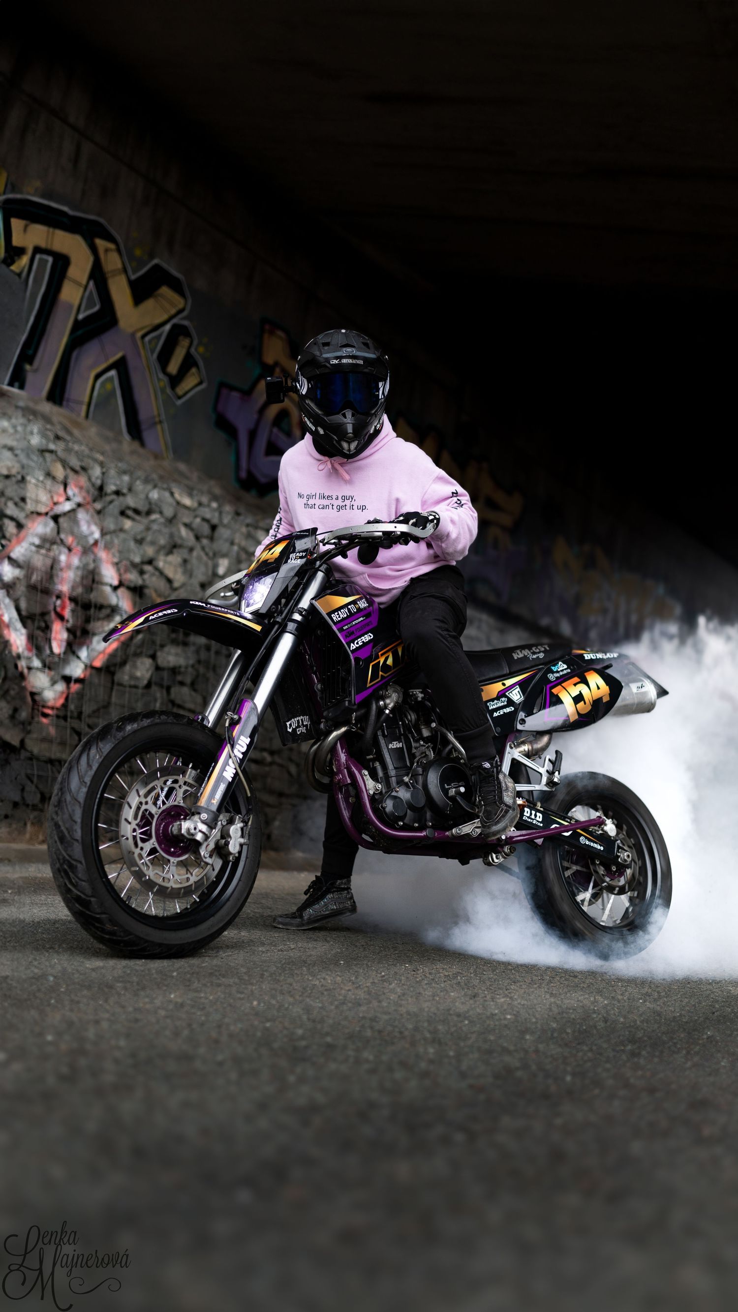 Motorcycle rider in pink shirt performing stunts with smoke effects in dark urban environment.