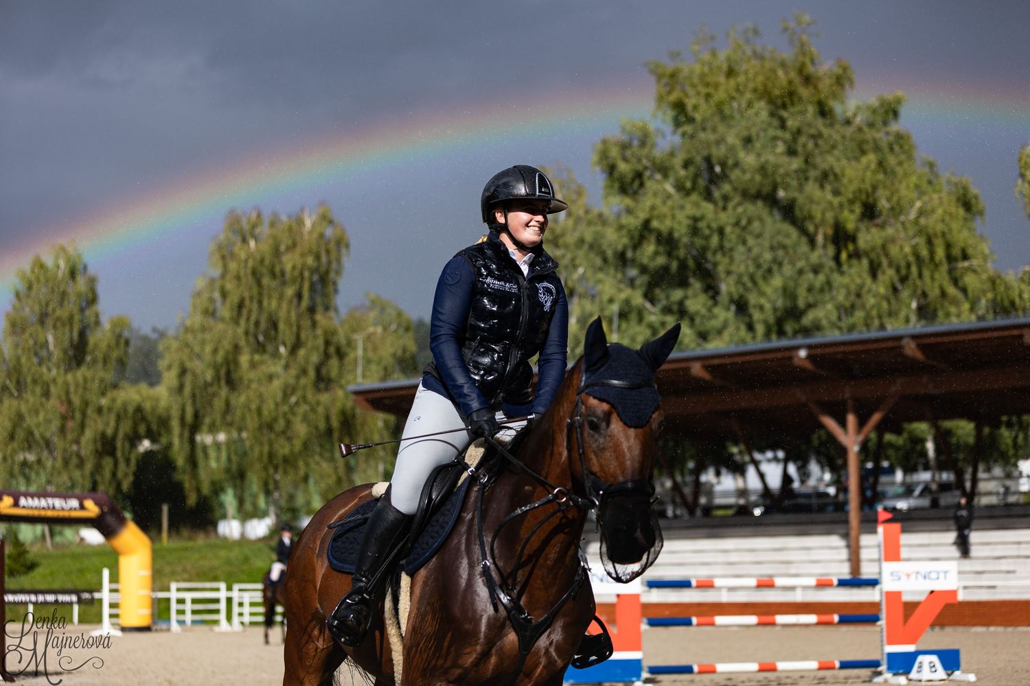A rider on horseback jumps over a colorful obstacle during an equestrian show jumping competition under a rainbow sky.