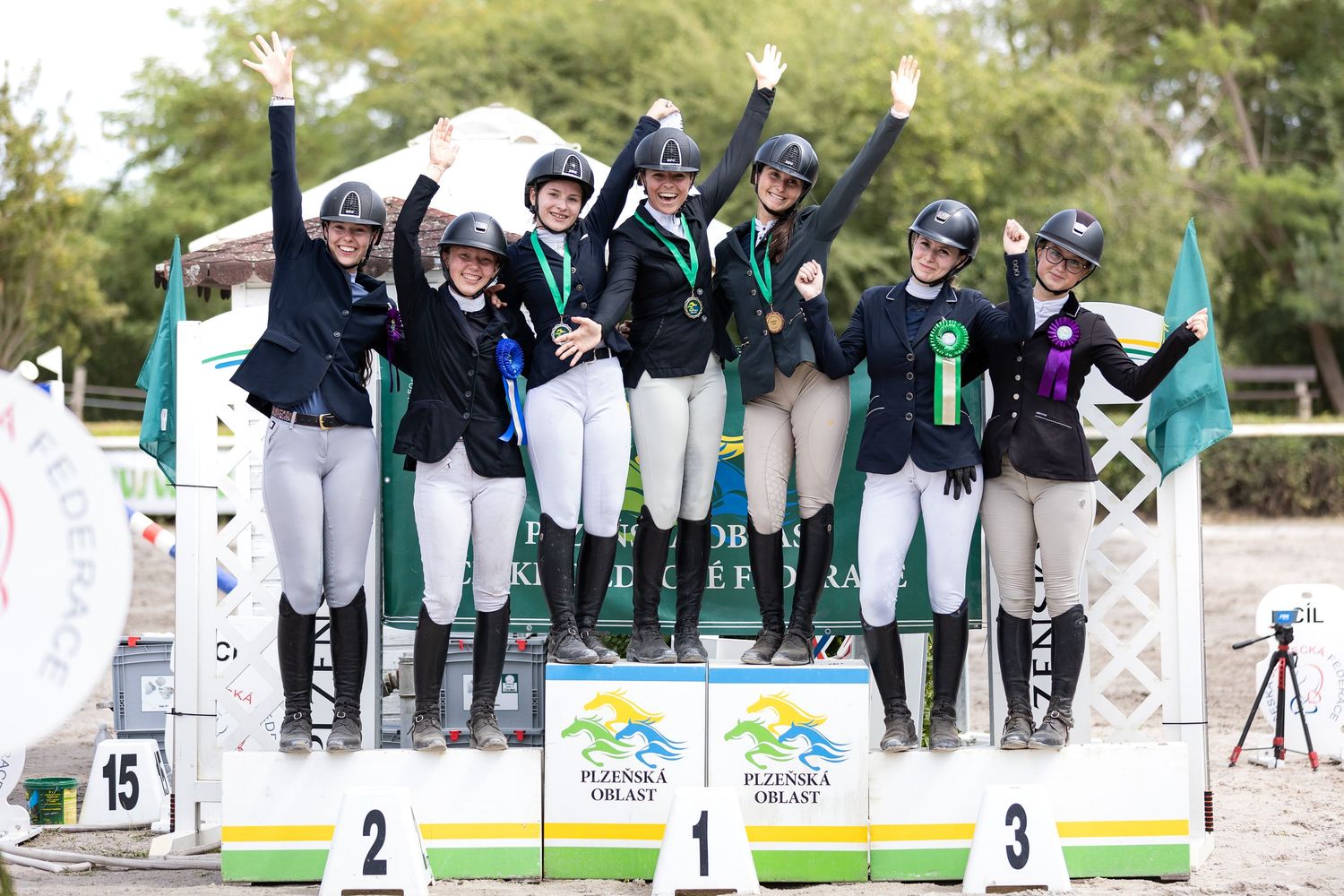 Equestrian team celebrates victory on winners podium at outdoor competition wearing matching uniforms and medals.