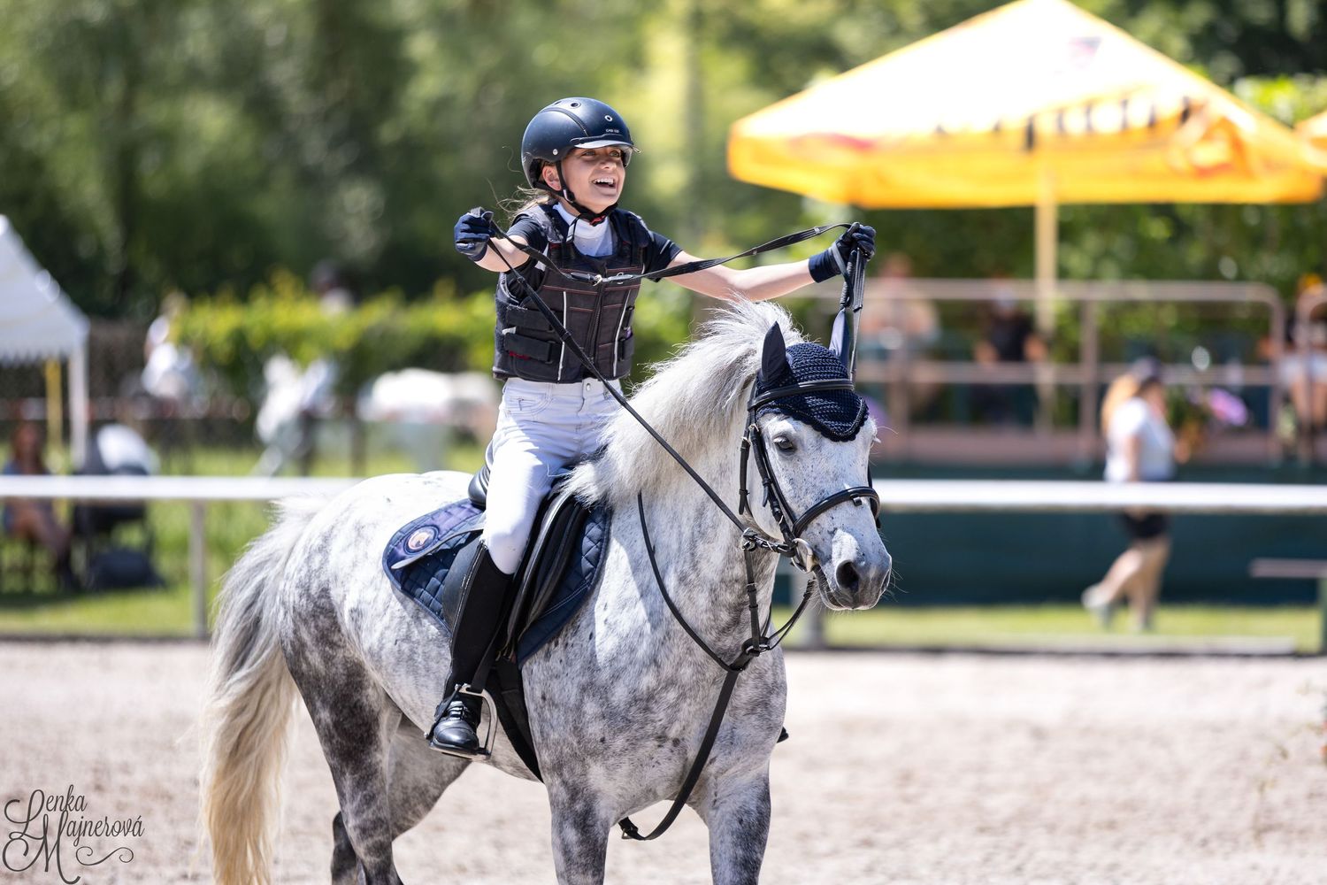 Equestrian rider in black helmet and vest gestures while riding a dappled grey horse in outdoor competition arena.
