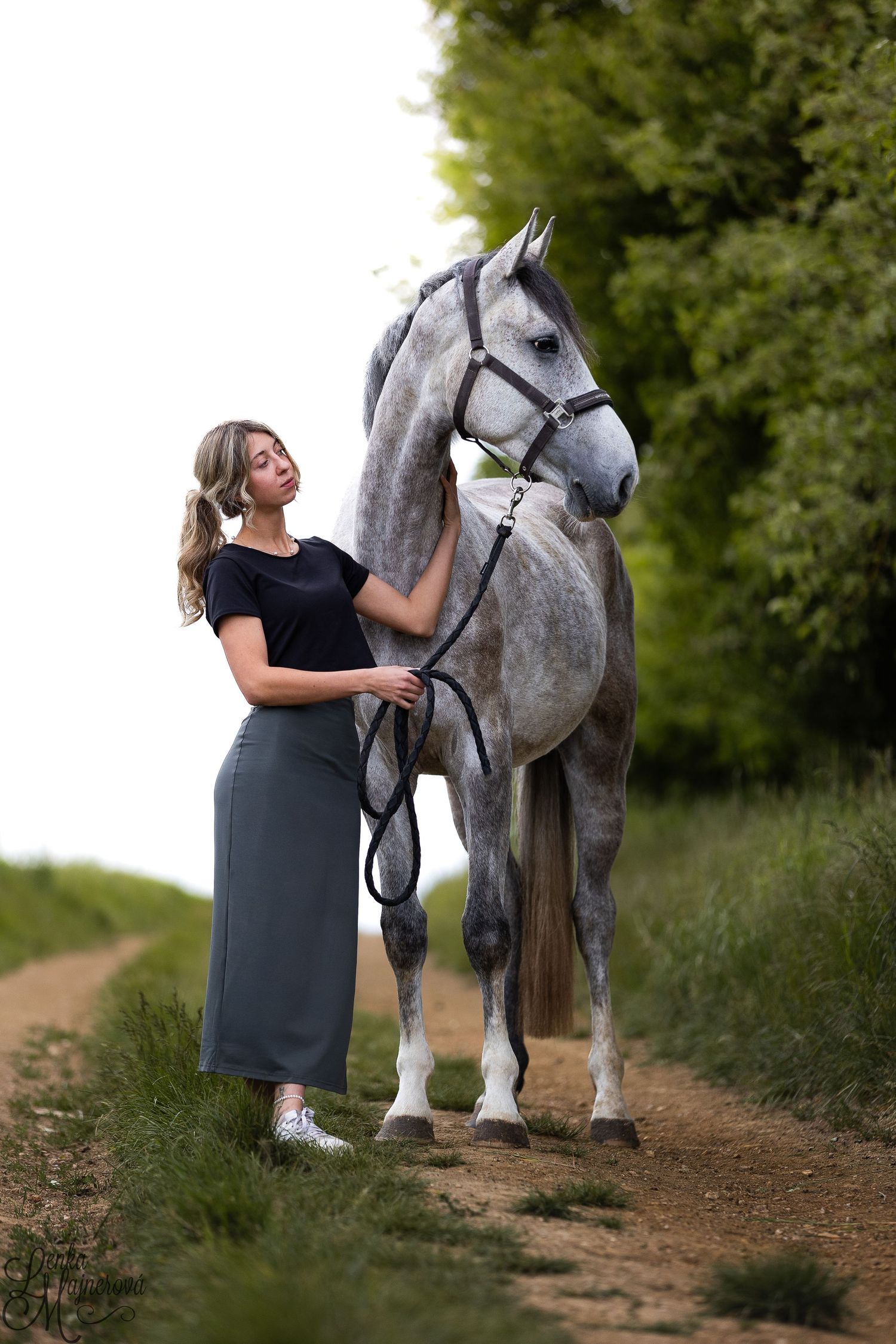 A pregnant woman in a gray dress poses with a majestic dappled gray horse on a rustic dirt path surrounded by greenery.