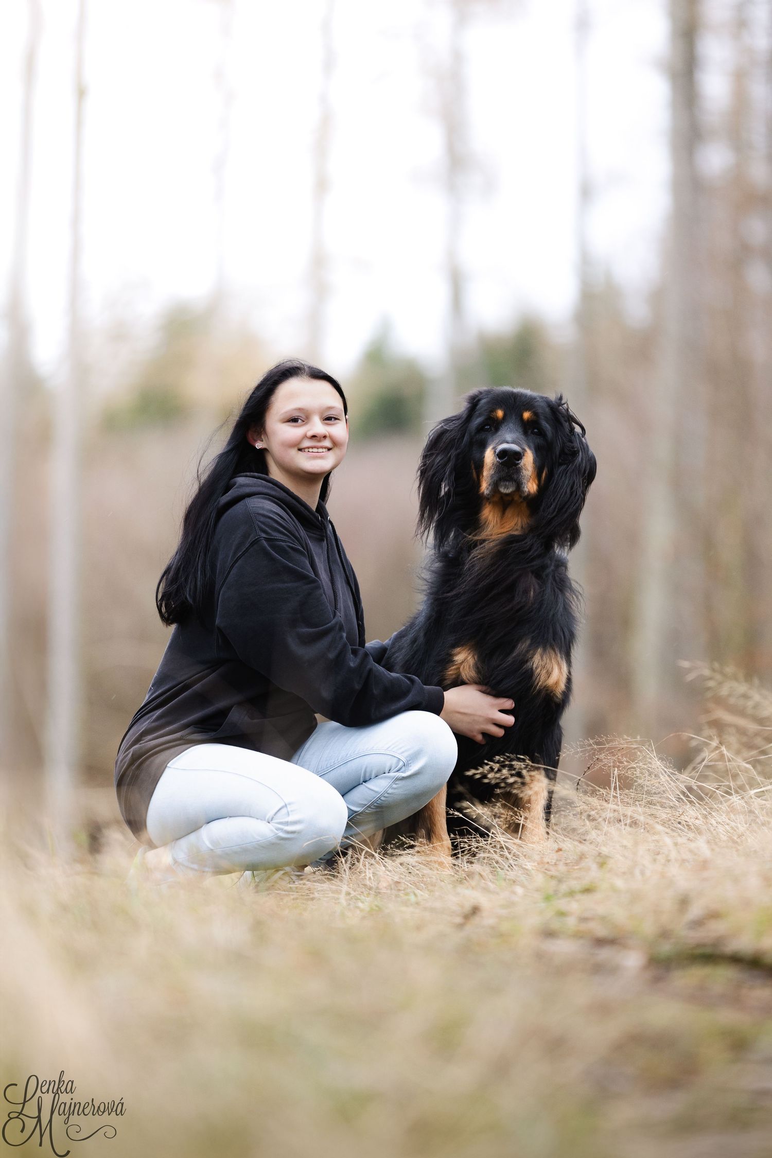 A person in a black hoodie crouches next to a large Hovawart dog in a natural outdoor setting with trees in background.