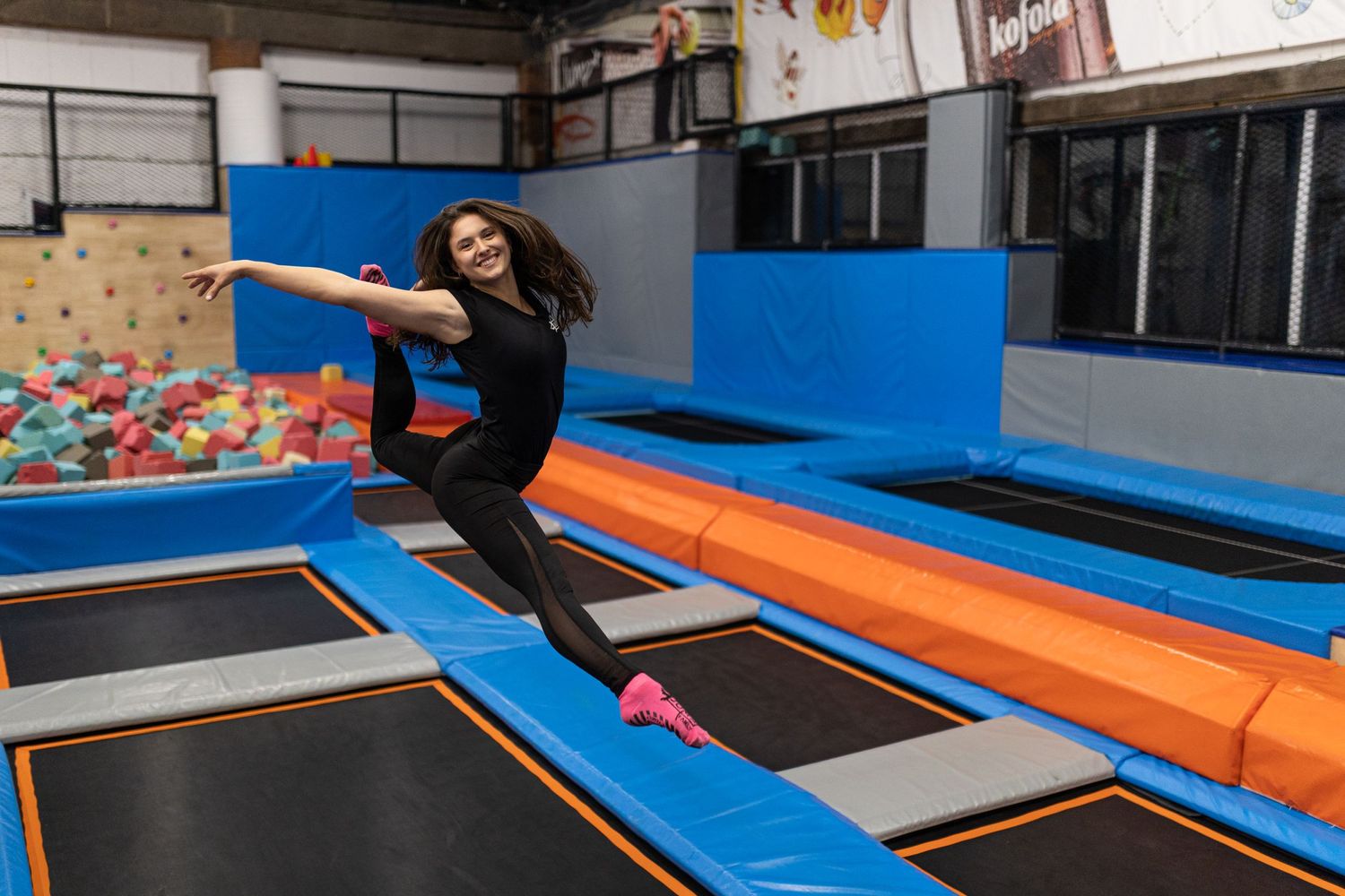Person jumping on a trampoline in an indoor recreation center with a foam pit and blue padding.