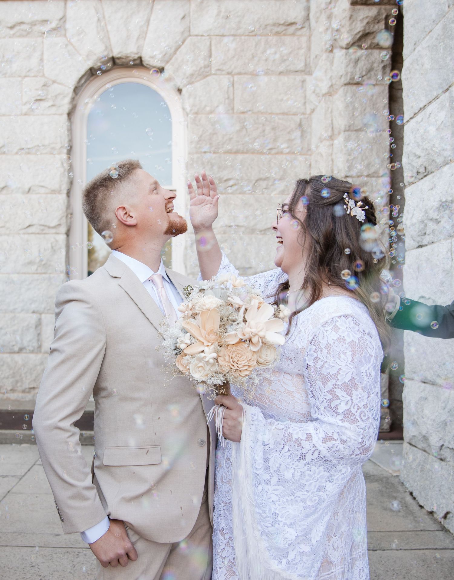A couple in wedding attire shares a playful moment against a white stone wall background.