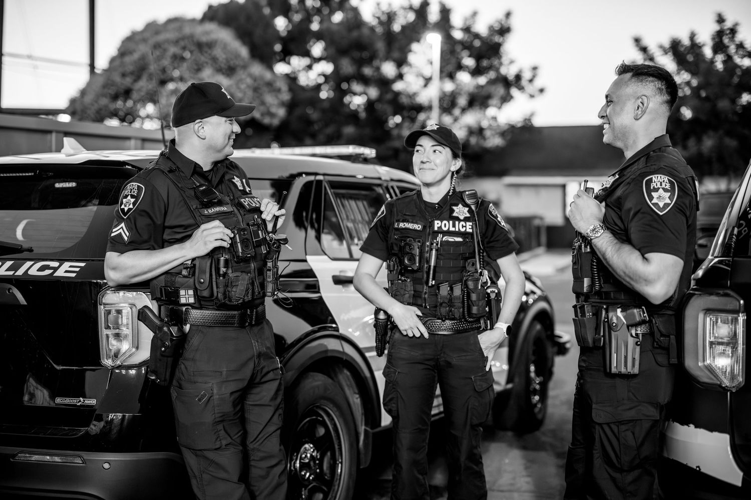 Police officers in tactical vests stand next to patrol vehicles during a nighttime operation in black and white.