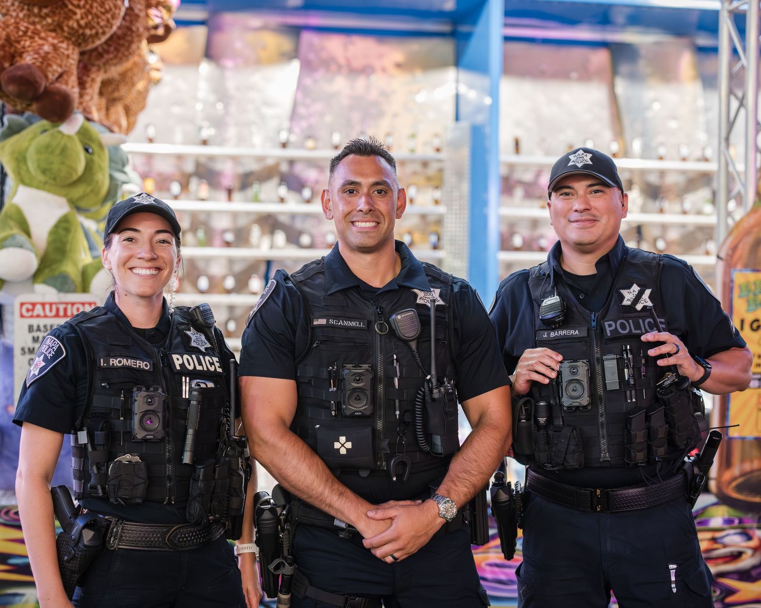 Three police officers in tactical vests stand together smiling at a community event.