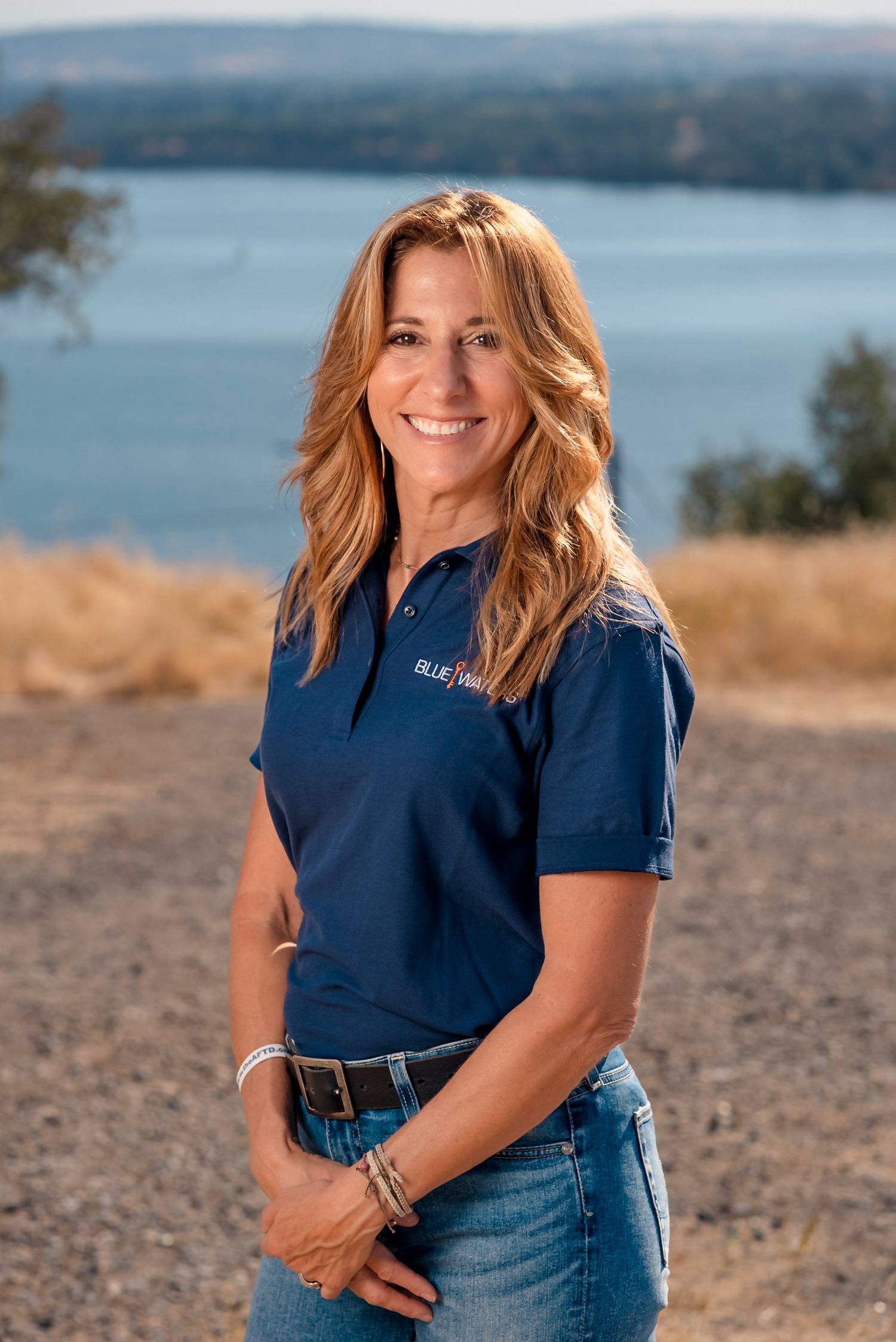 Woman in navy blue top and jeans poses outdoors against scenic waterfront backdrop on sunny day.