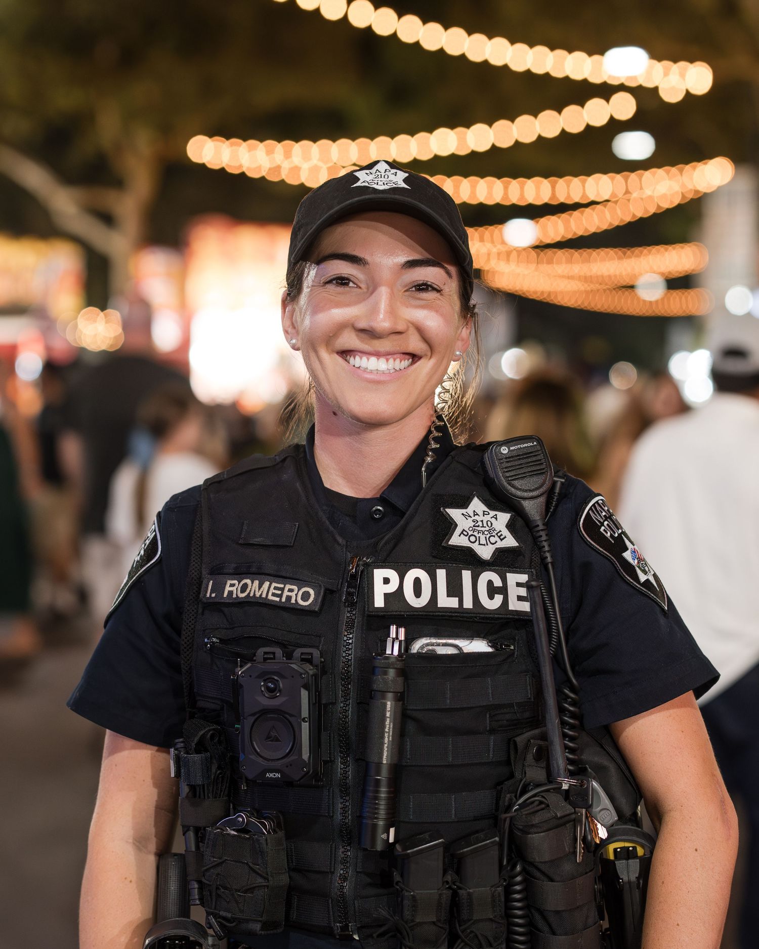 Police officer in tactical gear smiles while standing under decorative string lights at night.