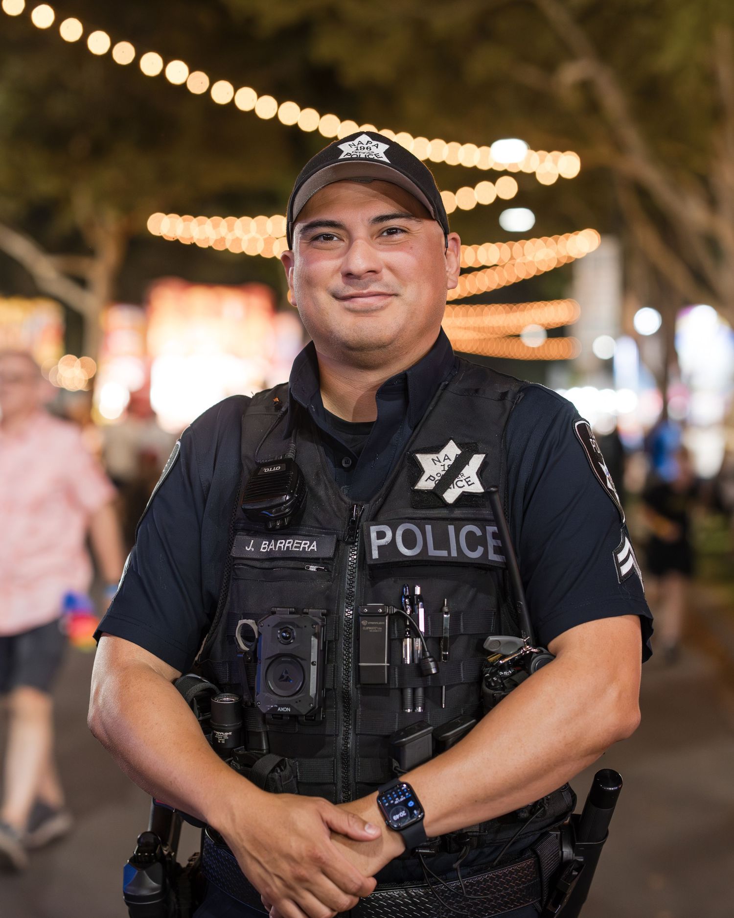 Police officer in tactical vest smiles at night event with string lights visible in background.