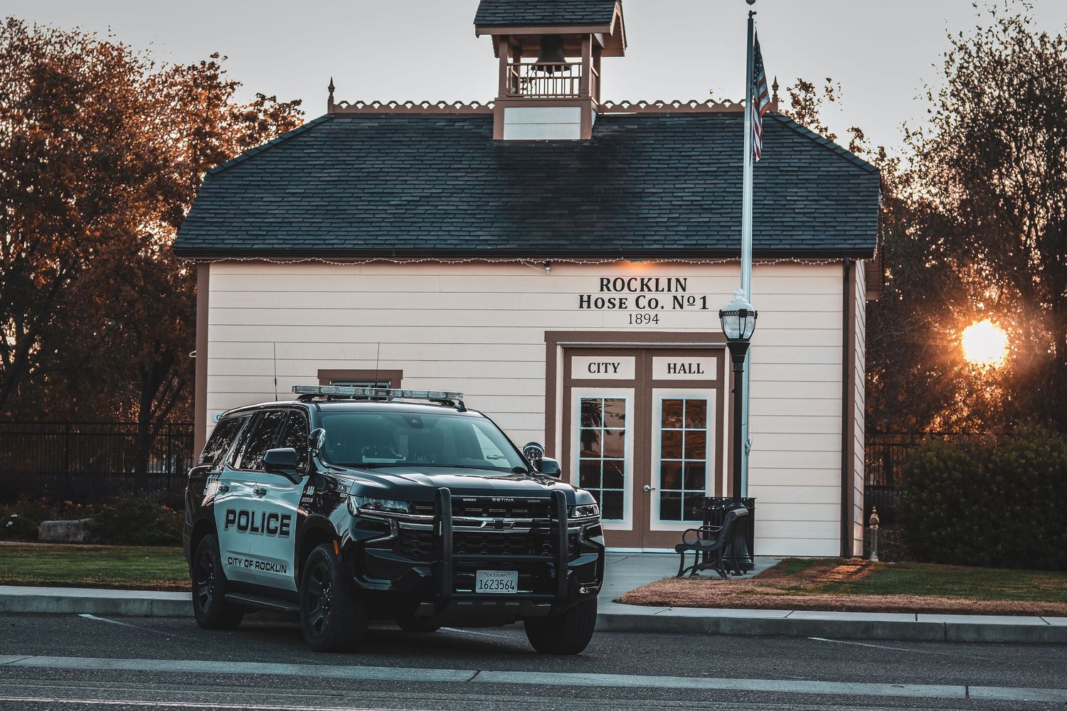 Police patrol car parked in front of a white historic schoolhouse building at sunset with glowing street lamp.