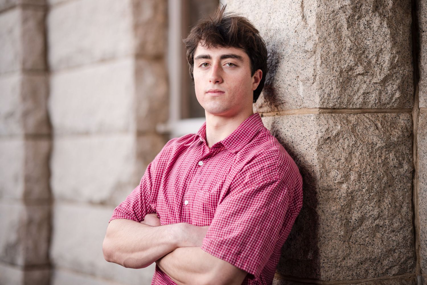 A person in a pink button-down shirt leans against a stone wall with arms crossed.