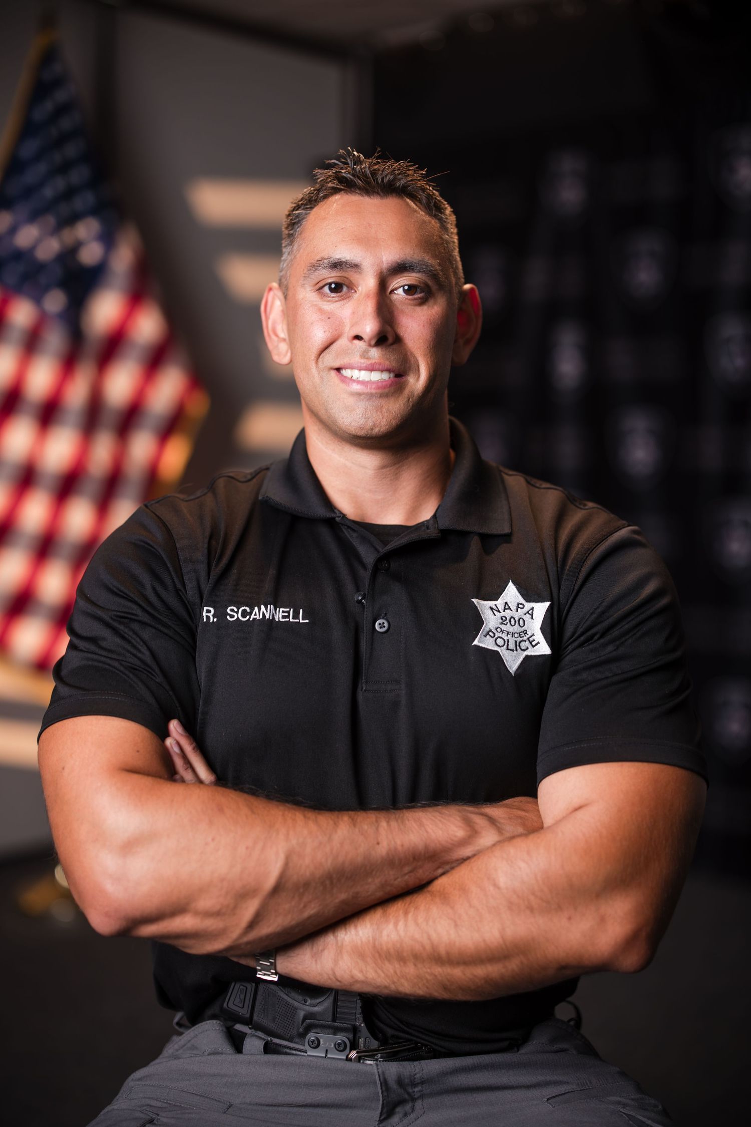 Law enforcement officer in black uniform poses against dark background with American flag for professional headshots.