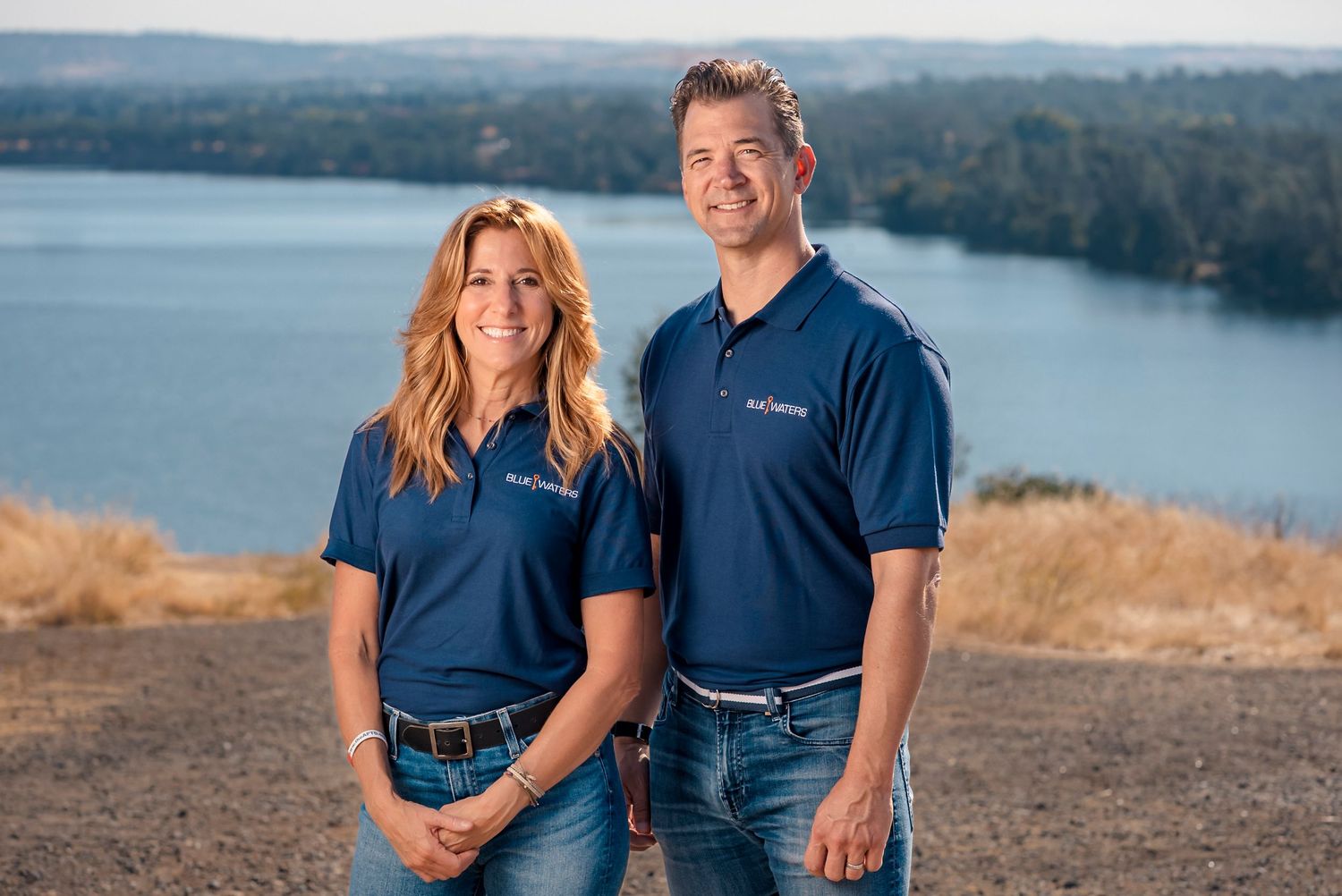 A couple in matching navy blue polo shirts poses together on a scenic hilltop overlooking a lake.