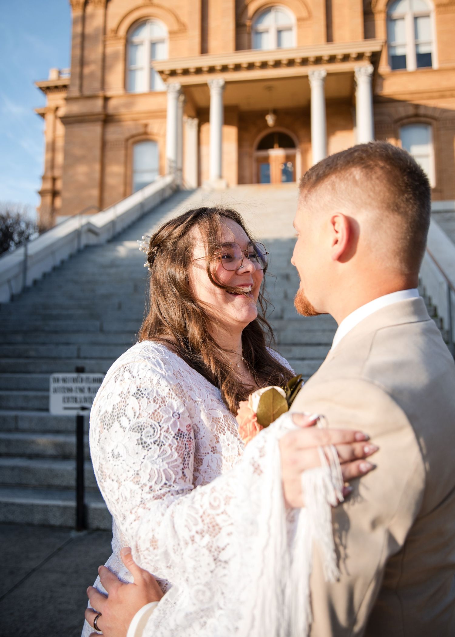 A bride and groom share a joyful moment on stone steps outside a historic building.