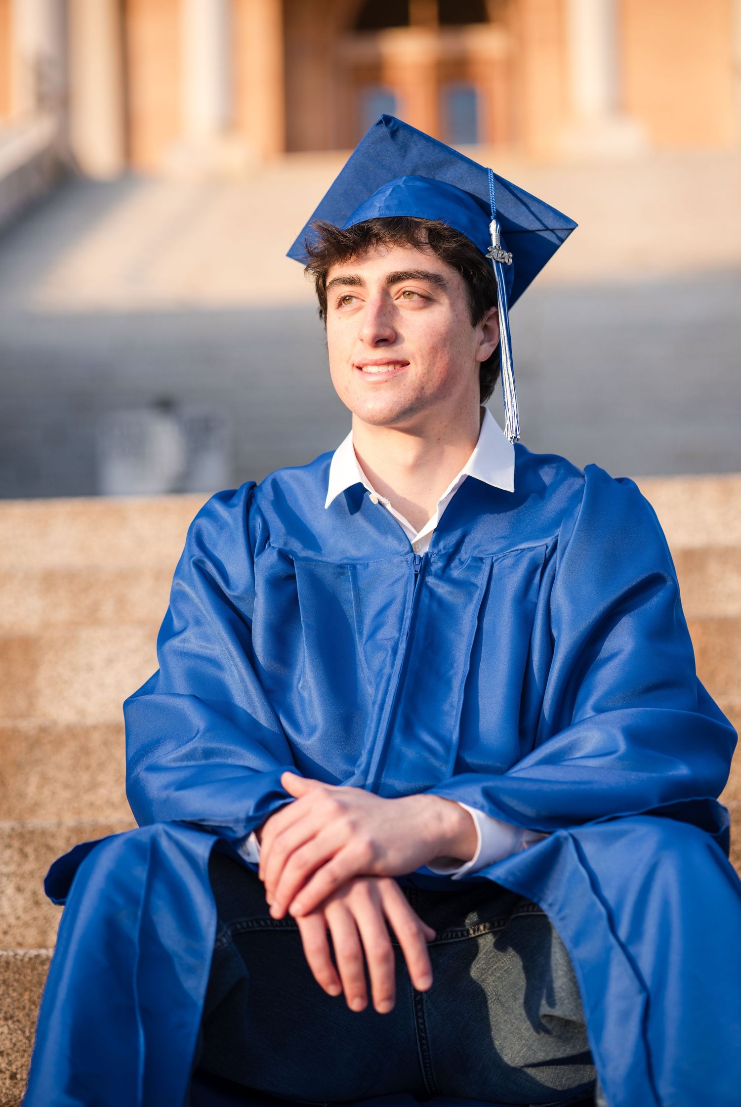 A graduate in a royal blue cap and gown sits outdoors during a photo session at sunset.