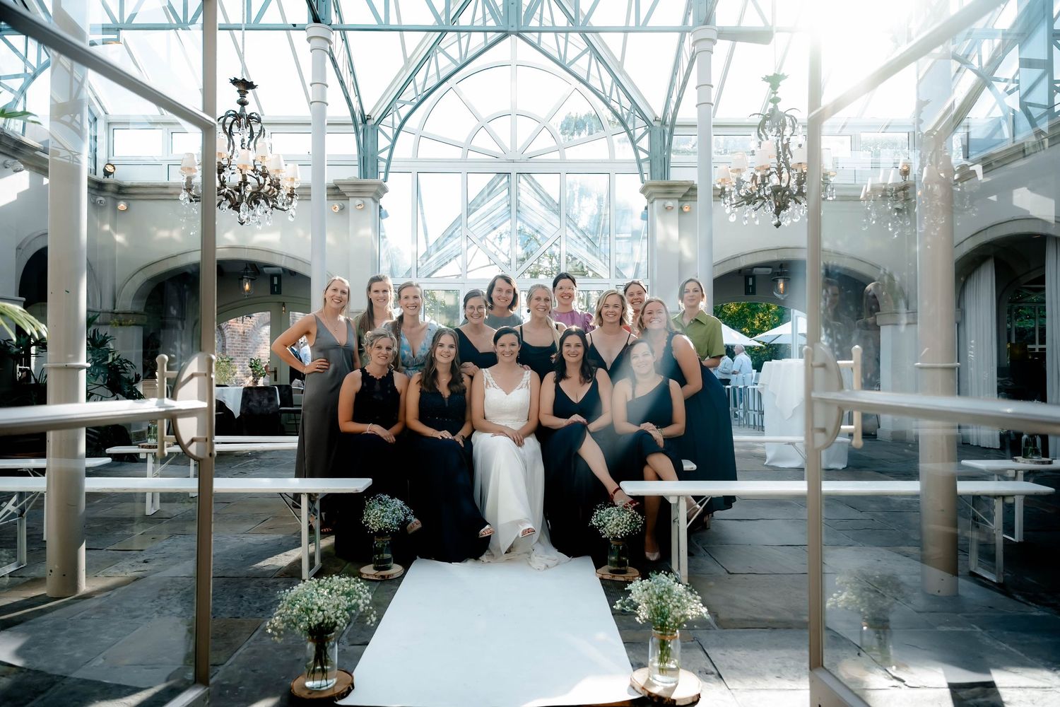 A wedding party poses together on elegant white benches inside a bright conservatory with ornate glass windows and chandeliers.
