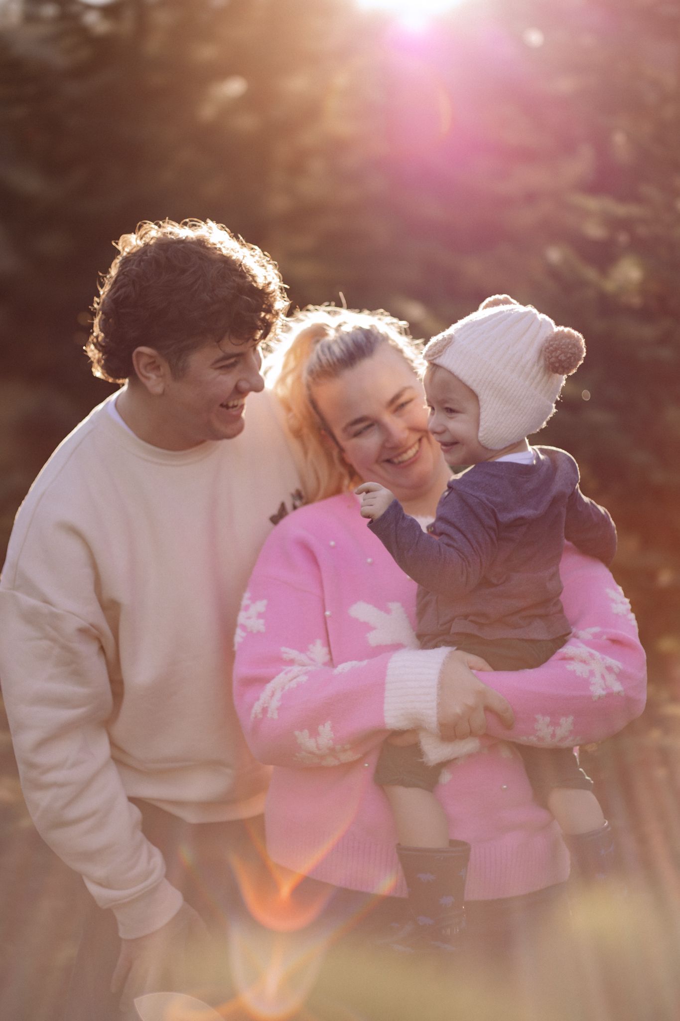 Young family with baby outdoors in golden sunlight, parents smiling, mom in pink sweater holding toddler in winter hat.