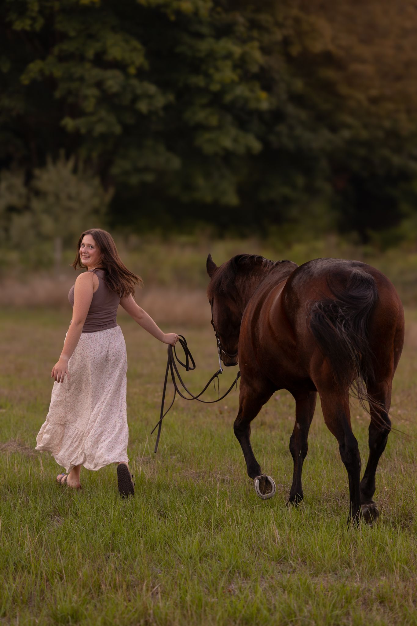 Woman in white skirt leads a bay horse across a green field with dark trees in the background.