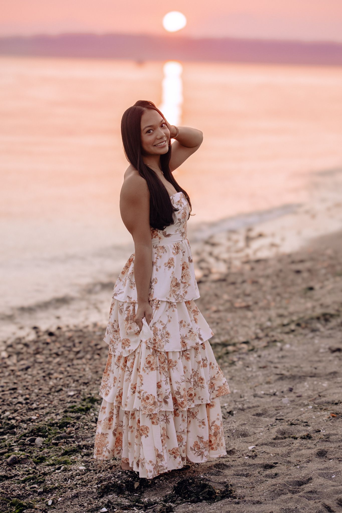 Woman in floral maxi dress posing on beach at sunset with pink sky and ocean reflection.
