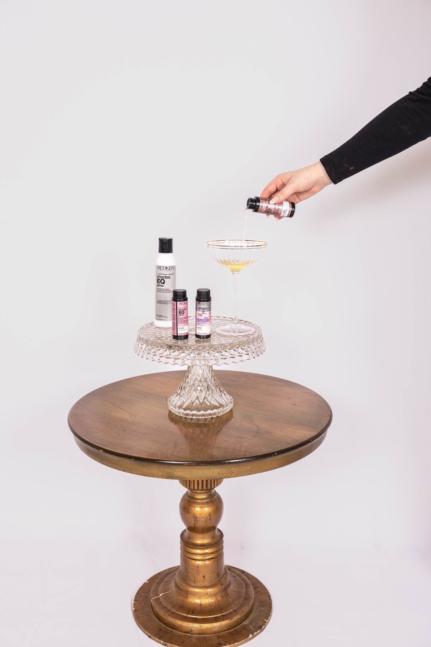 Hand pouring drops into a cocktail glass on a crystal cake stand with beauty bottles on a wooden pedestal table.