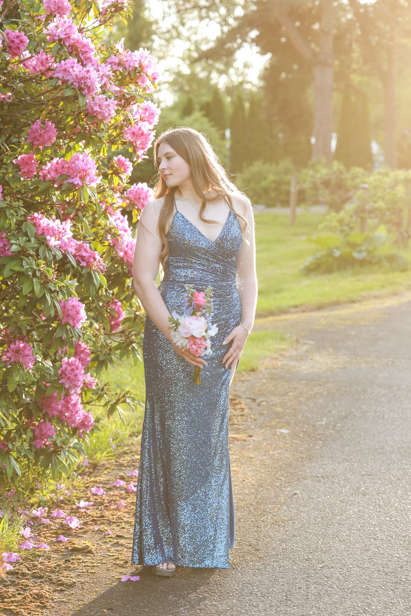 Young woman in silver sequin gown holds bouquet beside pink flowering bush in golden sunlight.