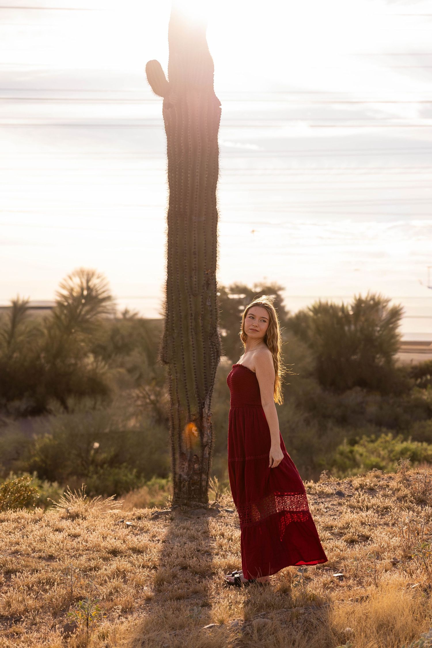 Young woman in red dress standing beside tall saguaro cactus at golden sunset in Arizona desert.