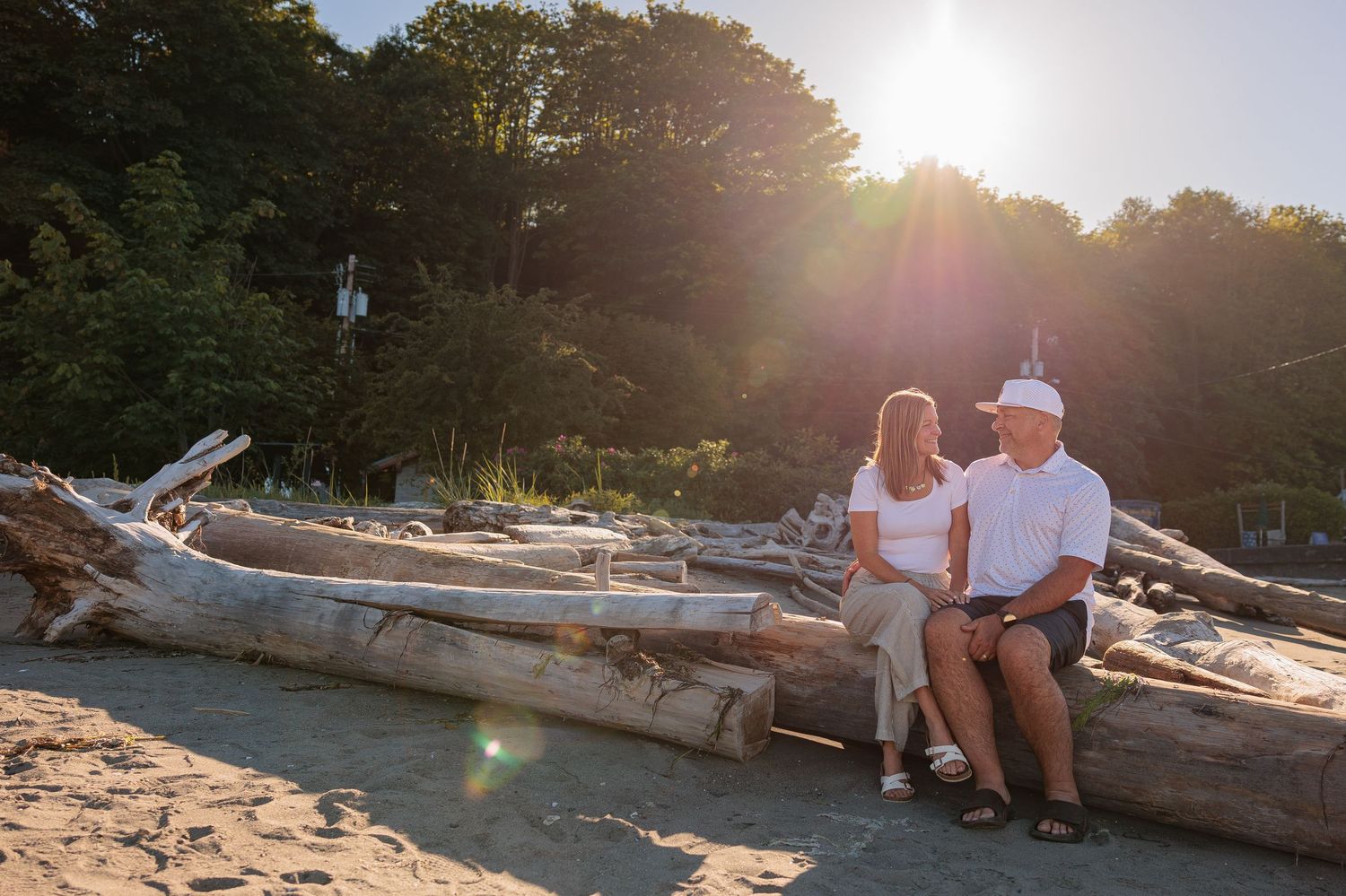 Couple sitting on driftwood logs at a sunny beach with lush green trees in the background at golden hour.