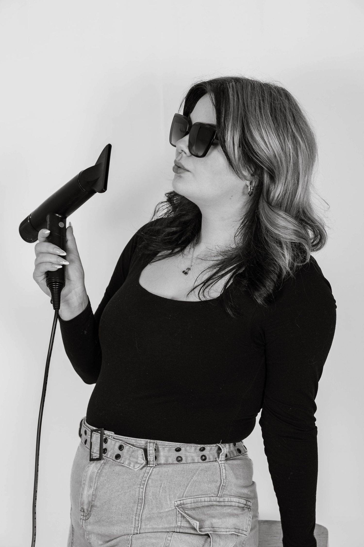 Woman in sunglasses holding a hair dryer, wearing black top and jeans, posing confidently in black and white photo.