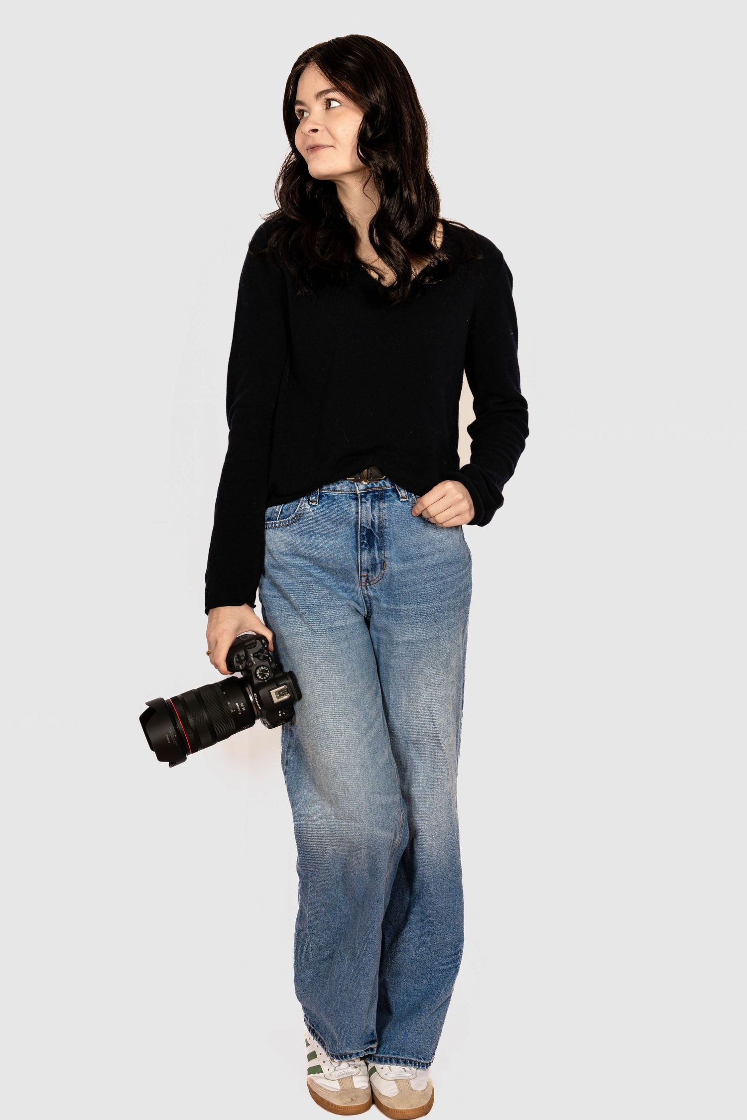 Young woman in black sweater and blue jeans holding a DSLR camera, standing against a light gray background.