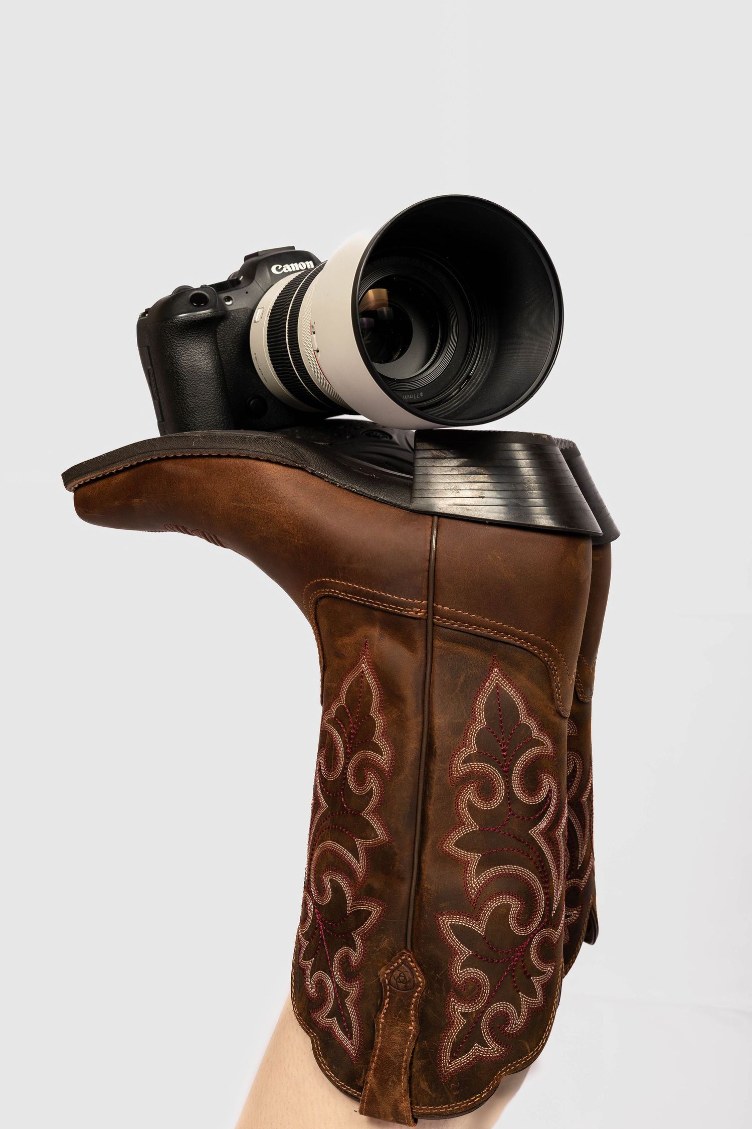 A Canon DSLR camera with telephoto lens balanced on a brown leather cowboy boot held up against white background.