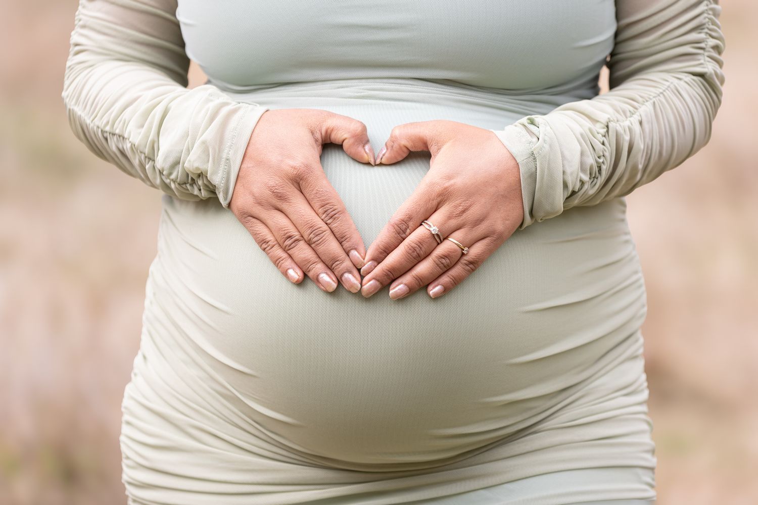 Pregnant woman forming heart shape with hands on baby bump wearing a sage green dress outdoors.