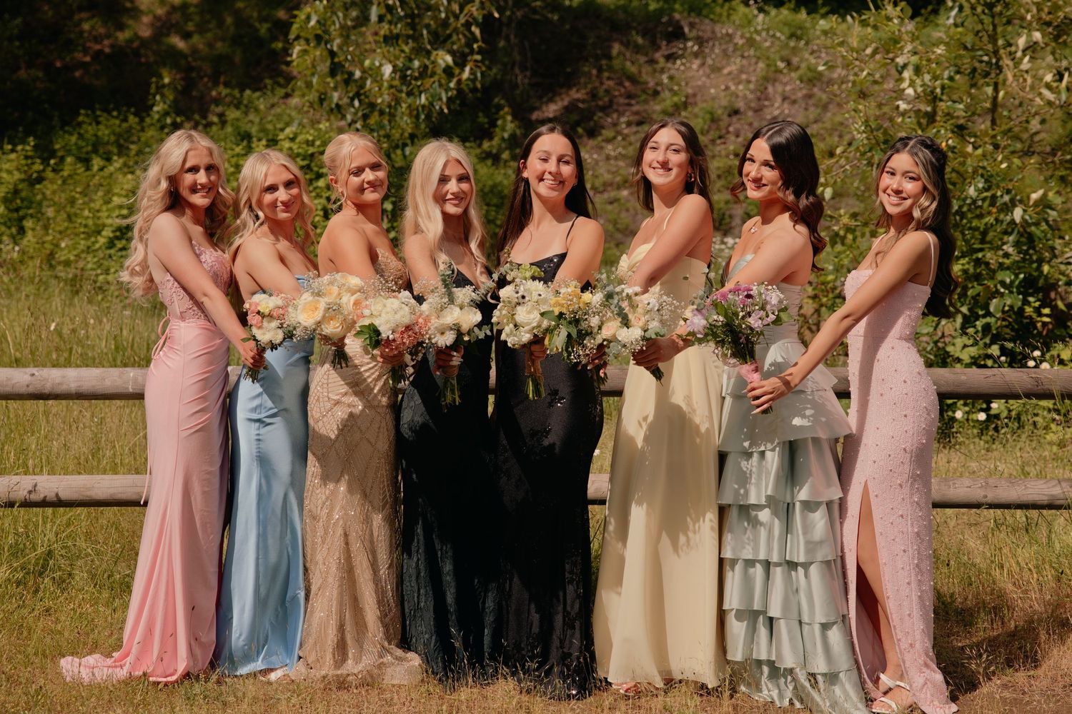 Seven bridesmaids in mismatched pastel gowns holding floral bouquets pose together outdoors near a wooden fence.