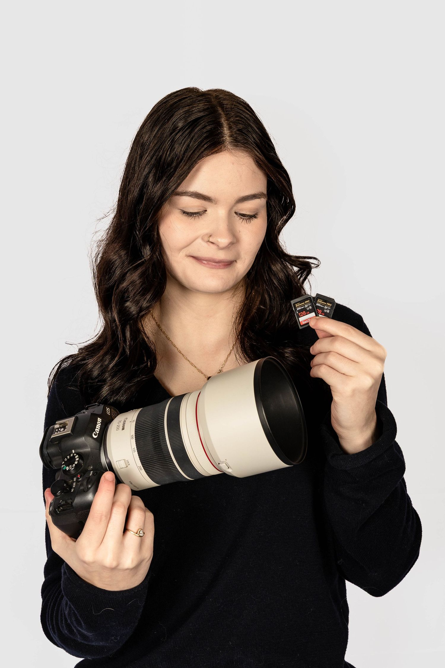 Young woman in black sweater holding a Canon DSLR camera with telephoto lens and memory card on white background.
