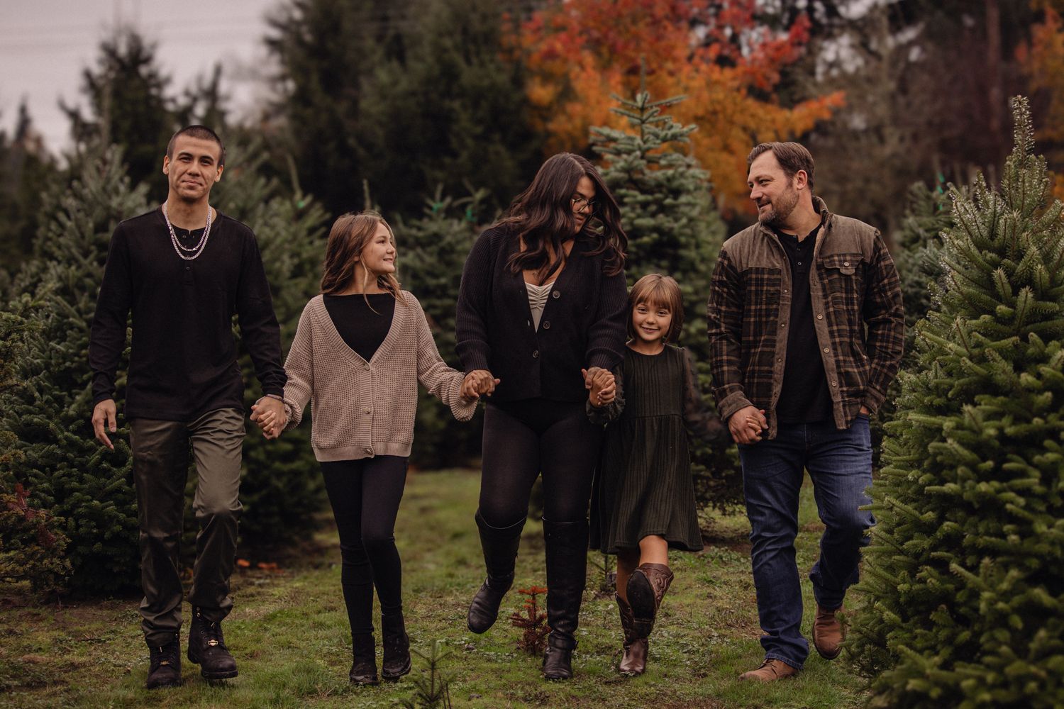 Family of five walking and holding hands at a Christmas tree farm with colorful autumn foliage in background.