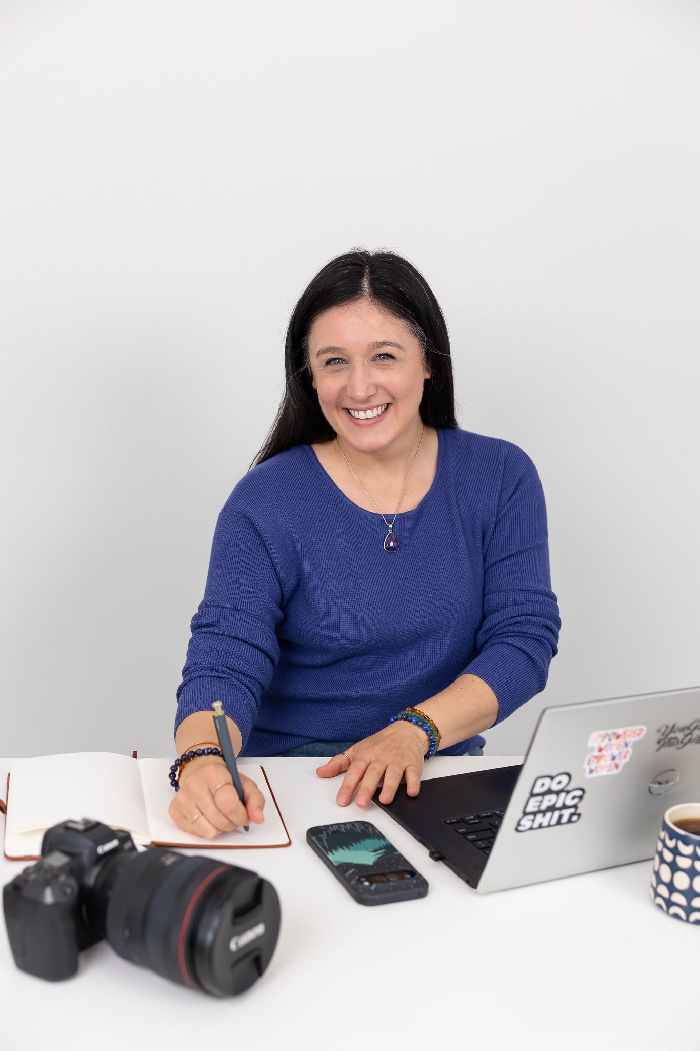 woman branding photographer sitting at desk with laptop, camera and writing in journal for headshot smiling at camera in Auburn, WA studio