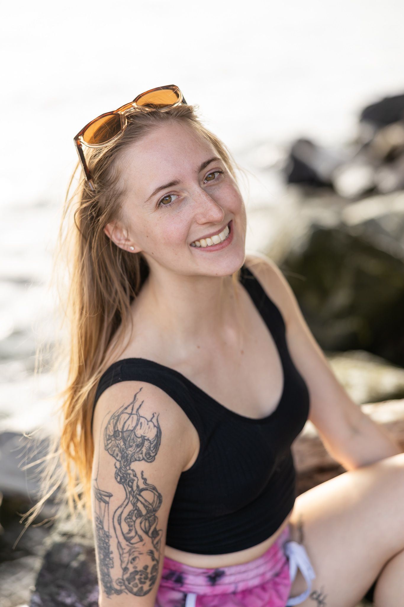 youngest blond woman sitting on beach for lifestyle headshot smiling at camera, sunsetting behind with sunglass on head