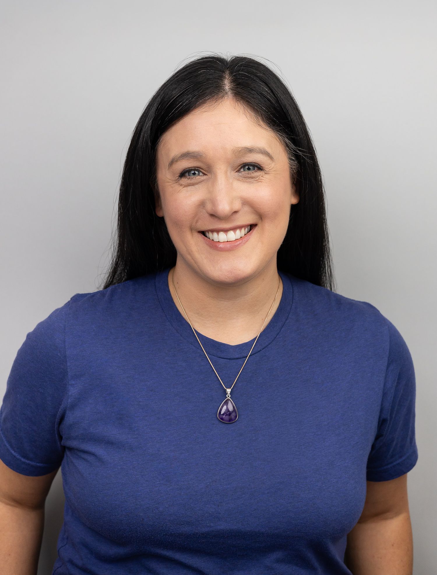 woman branding photographer wearing blue shirt on gray backdrop smiling for headshot in Seattle, WA wearing amethyst necklace