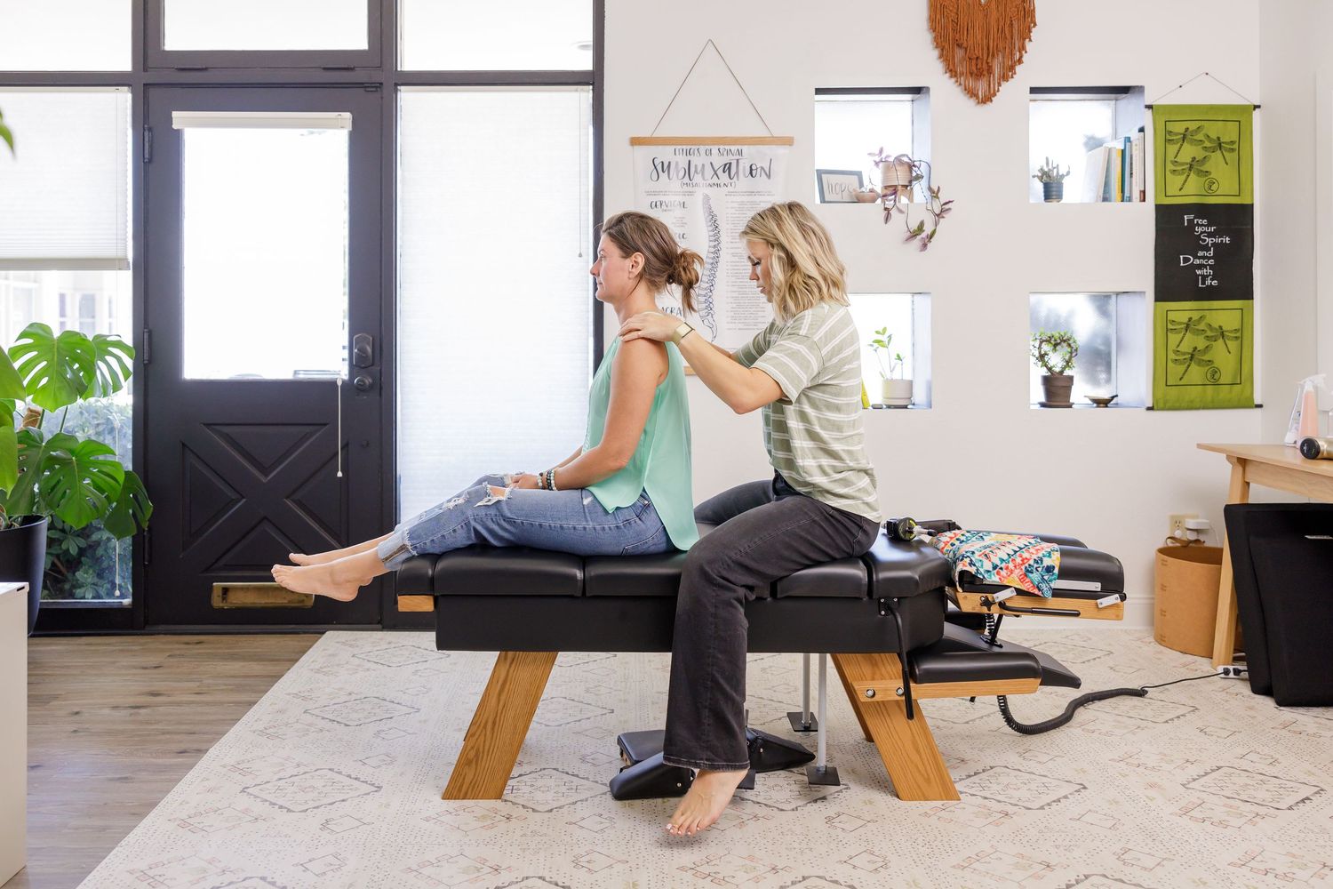 Woman chiropractor in Tacoma, WA sitting on table manually palpating shoulders of female client in office for lifestyle branding headshot