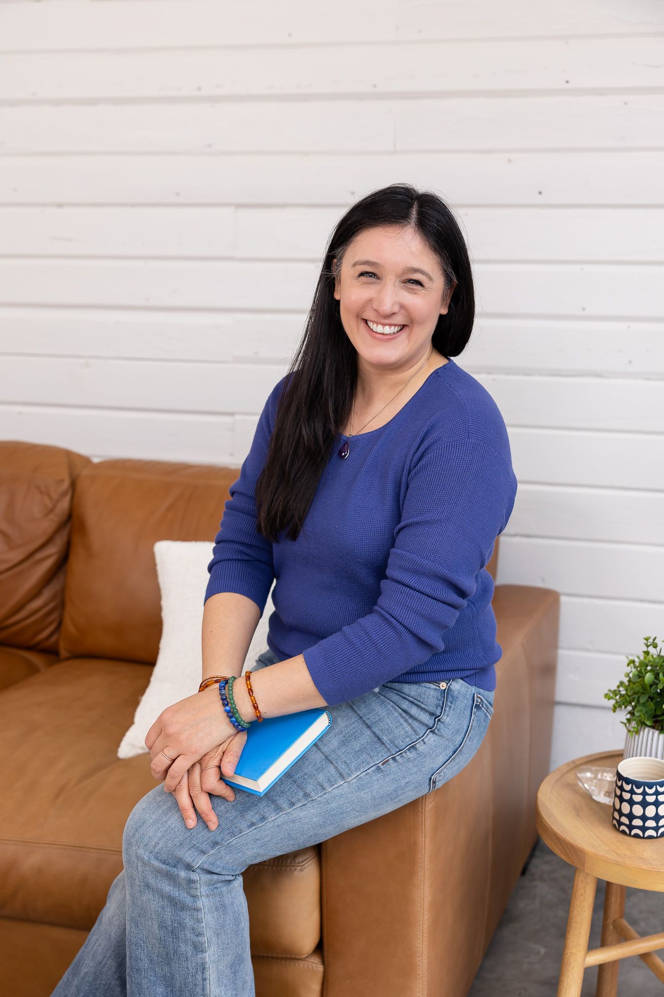 woman sitting on couch arm wearing blue sweater with book on lap laughing looking at camera for lifestyle headshot in Auburn, WA studio