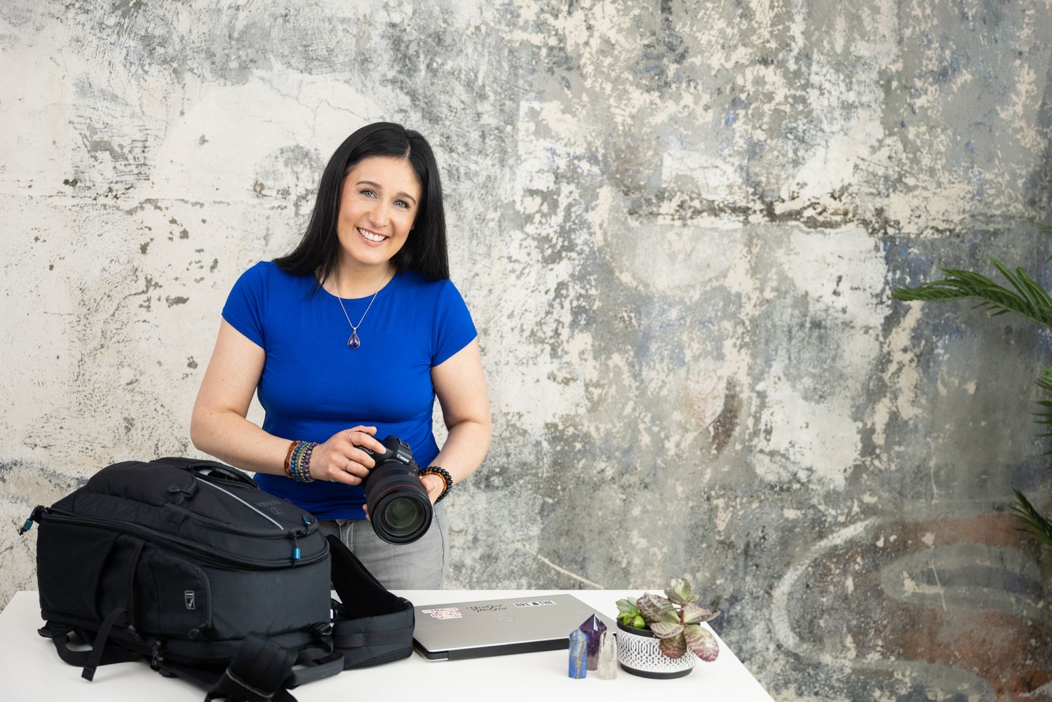 women branding photographer hold camera, camera bag, laptop on desk for lifestyle photo smiling at camera in studio in Seattle, WA