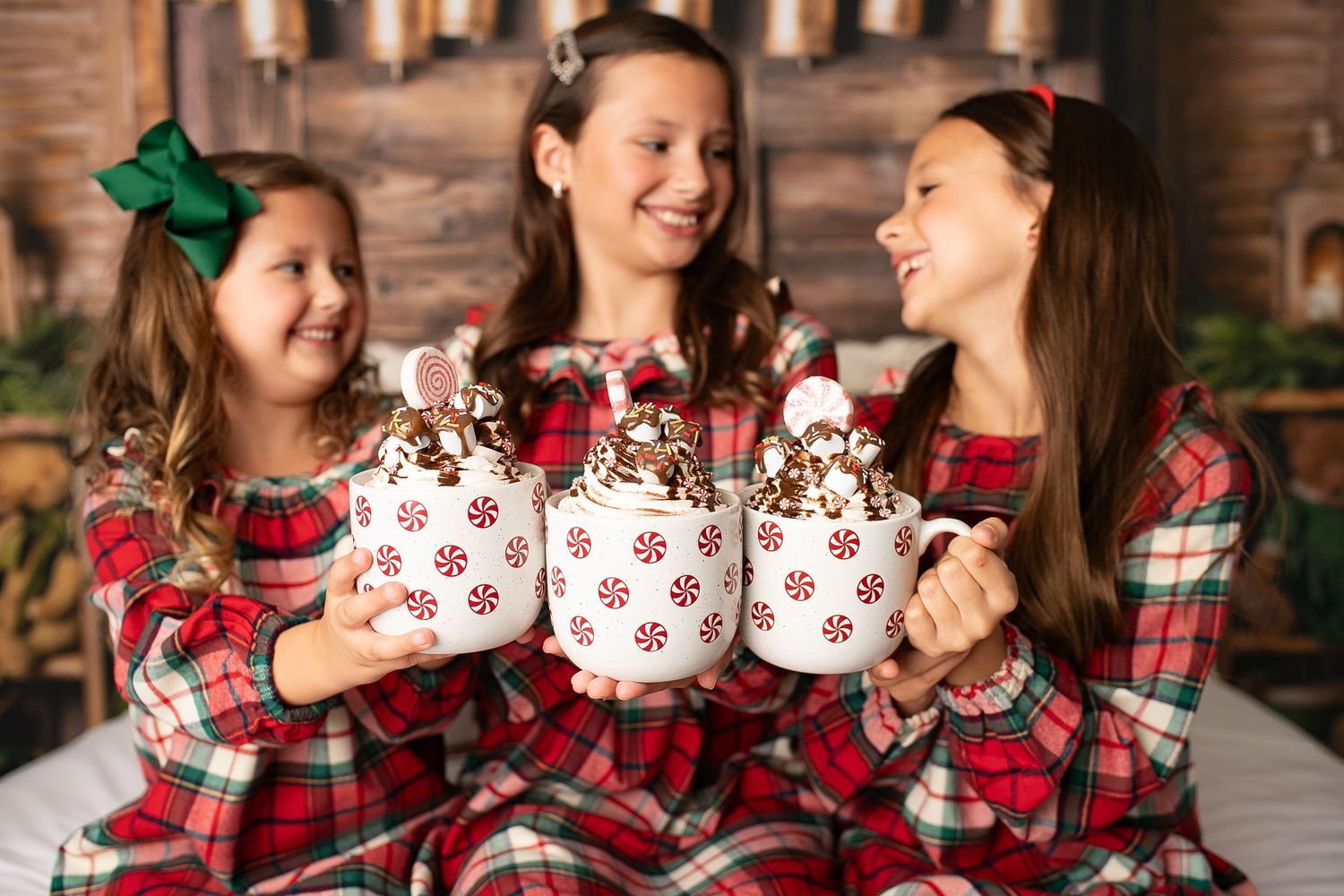 Three girls in matching red plaid Christmas pajamas holding festive mugs of hot cocoa with peppermint decorations.