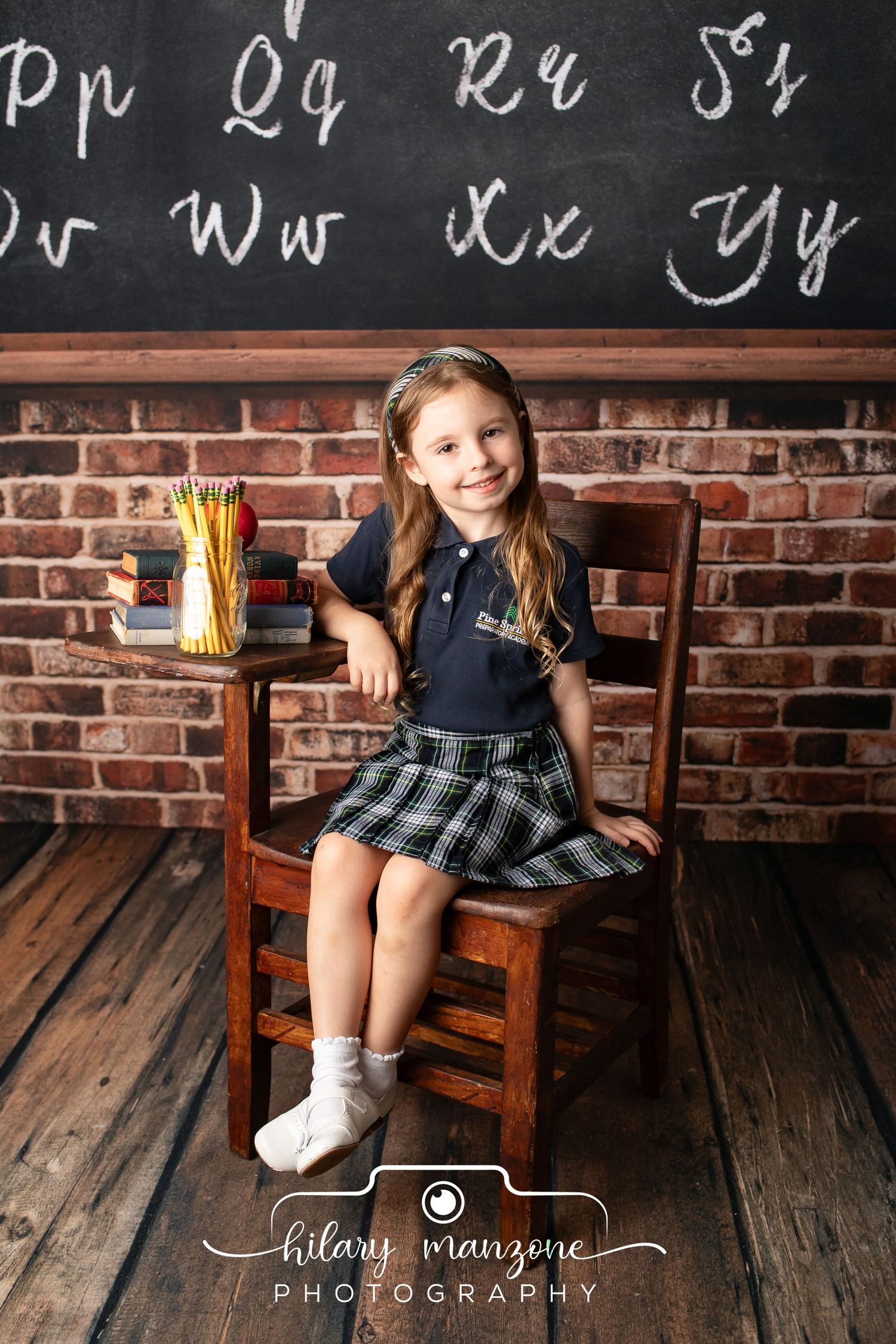 Young girl in school uniform smiles at desk with chalkboard alphabet backdrop in professional photo by Hilary Manzone.