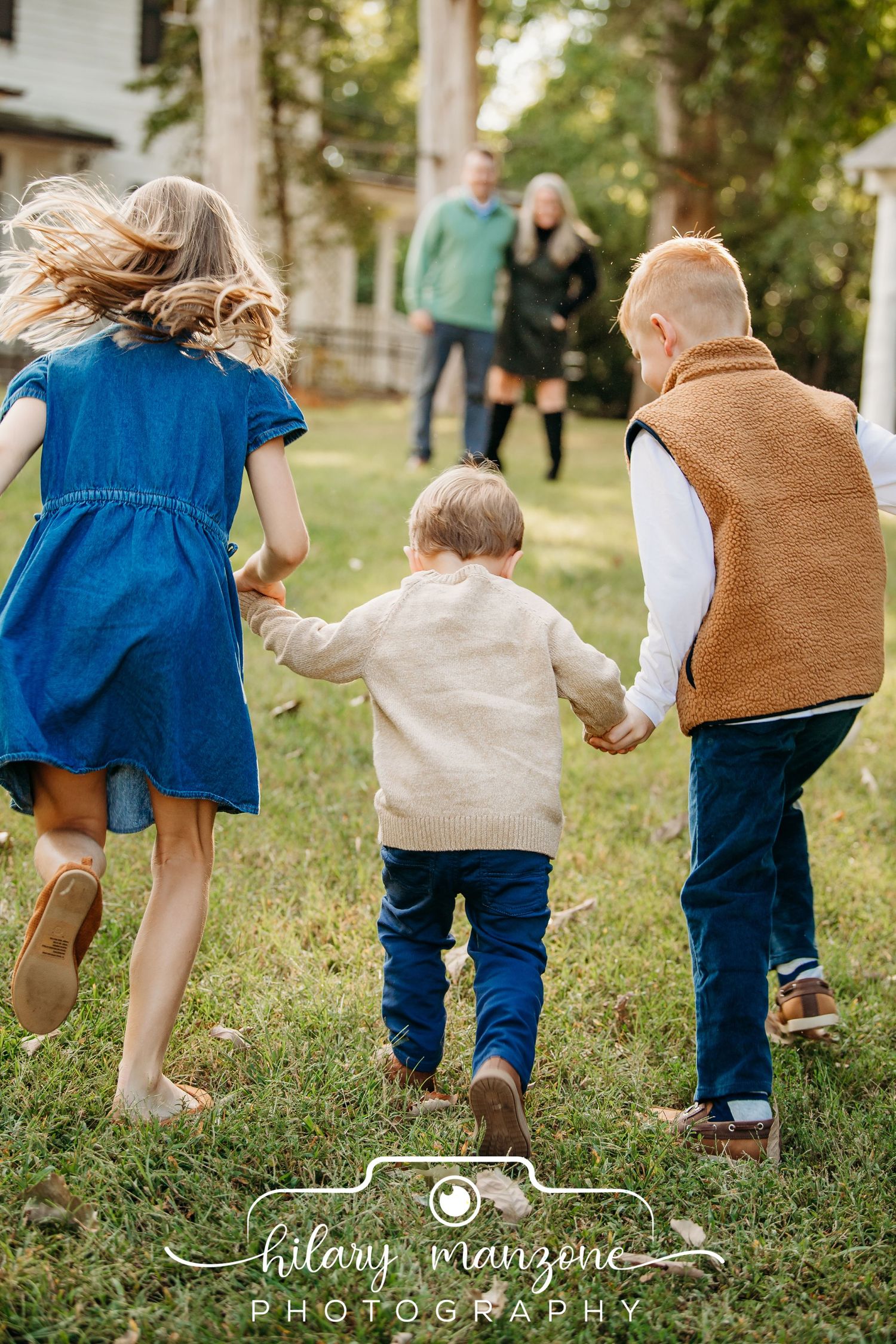 Three children run hand-in-hand toward their parents across a grassy lawn in a warm family photography session.