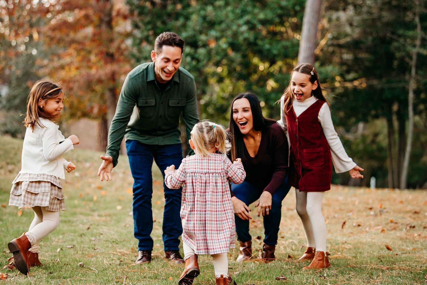 Happy family of five playing outdoors in autumn park, parents and three young daughters laughing together.